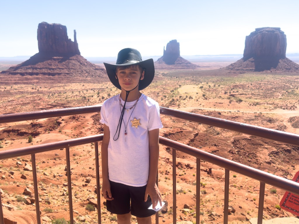 Bastion in a cowboy hat and sheriff badge at a railing, with Monument Valley buttes behind him under a blue sky.