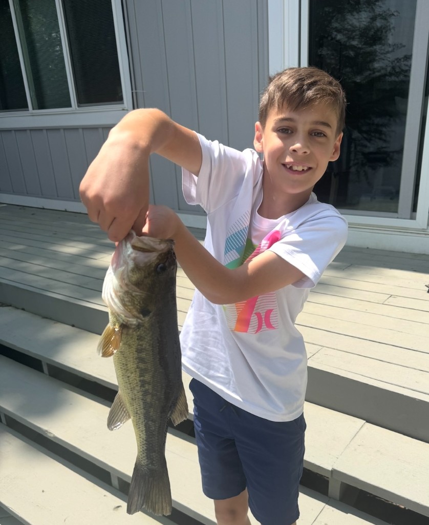 Bastion smiling on a deck, holding a large largemouth bass by the jaw.