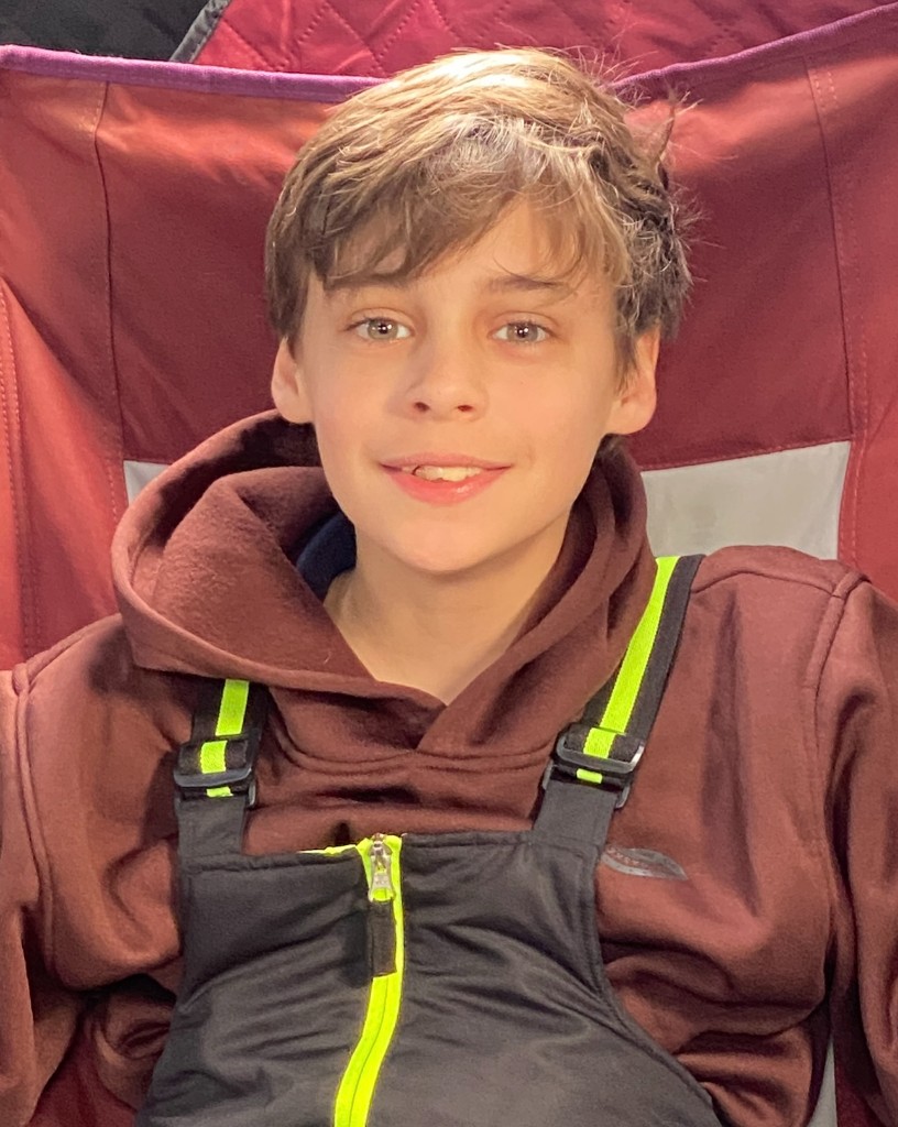 Bastion in winter bibs and a hoodie, sitting in a folding chair inside an ice-fishing shelter, smiling at the camera.