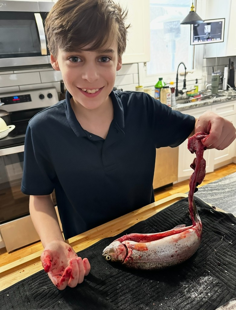 Bastion in a navy polo cleaning a gutted rainbow trout on a cutting board in the kitchen.