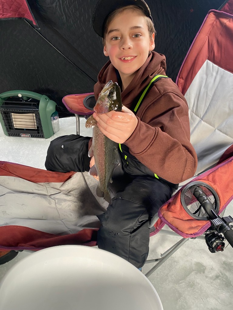 Bastion smiling in an ice fishing tent, holding a rainbow trout vertically with both hands.