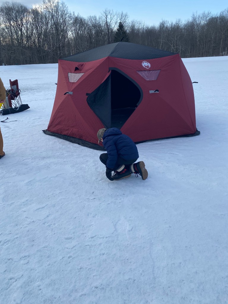 Inside an ice fishing tent: Bastion in winter bibs sits in a chair by a hole in the ice, with a yellow auger and propane heaters visible.