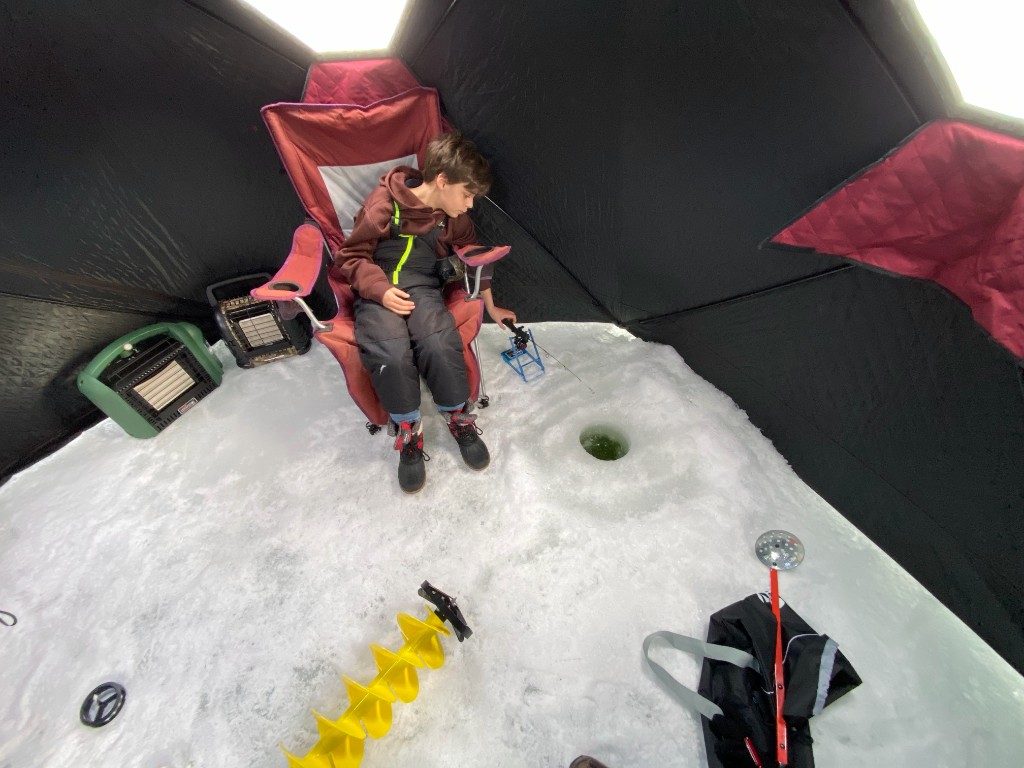 Bastion kneeling in snow in front of a large maroon and black ice fishing tent on a frozen lake, trees on the horizon.