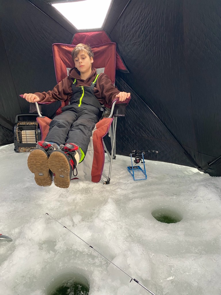 Bastion in winter gear sitting in a folding chair between two drilled ice holes with fishing rods on stands.