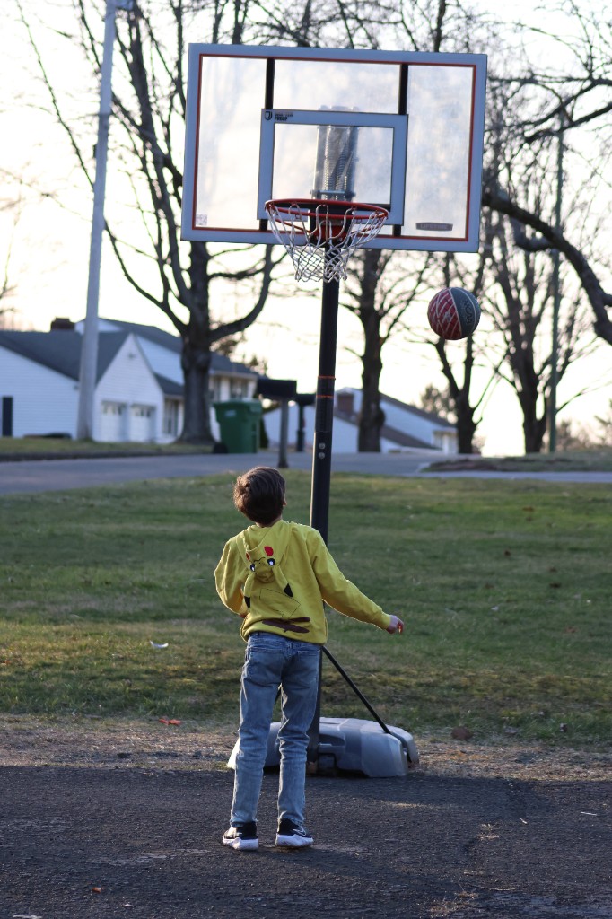 Bastion in a yellow Pikachu hoodie shoots a basketball toward a portable hoop on the driveway at golden hour.