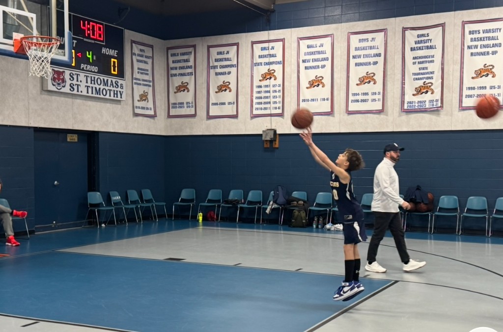 Bastion in a navy number 0 jersey jumps to release a shot in a school gymnasium with scoreboard and banners behind him.